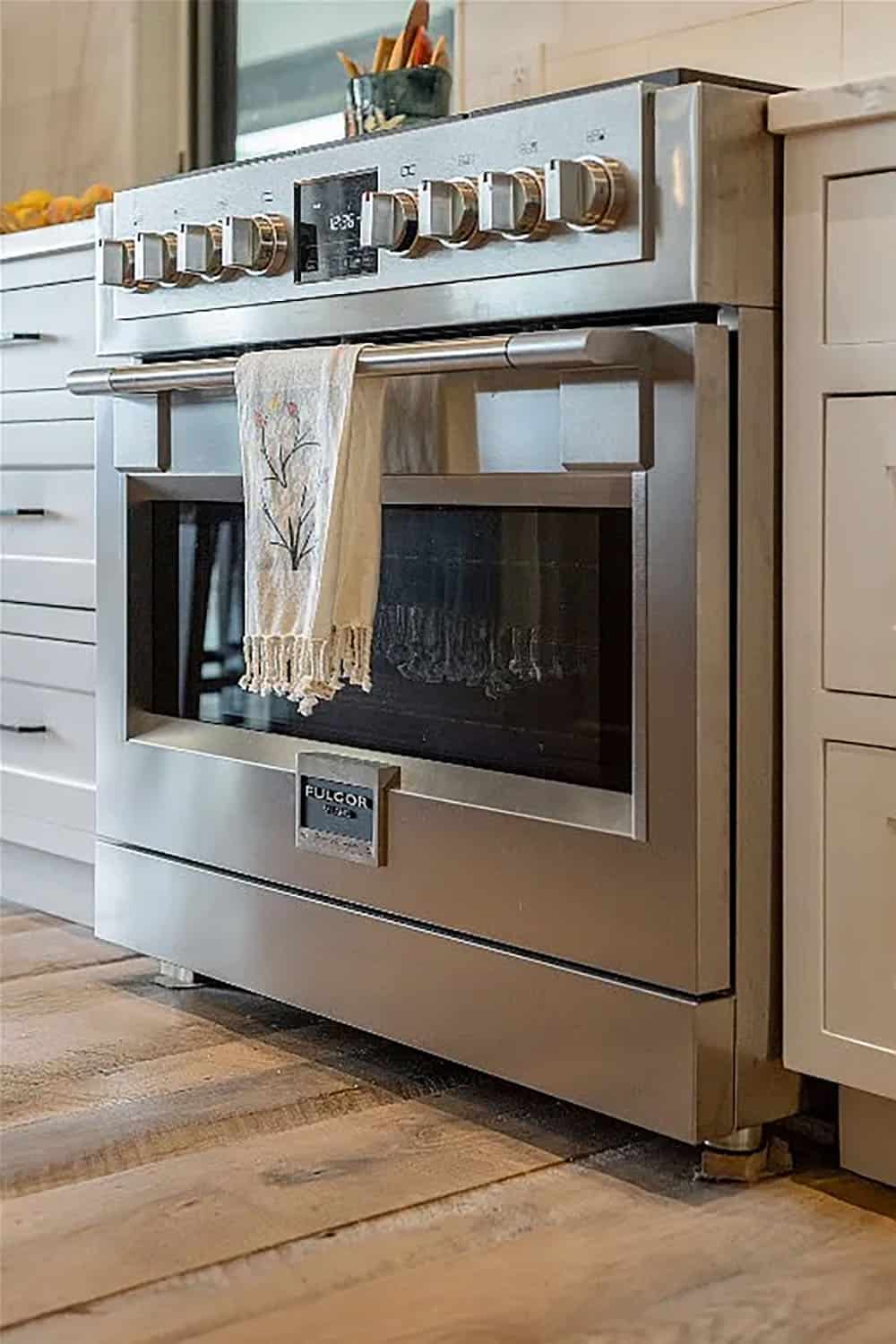 Close-up of Fulgor Milano stainless steel pro range with brass knobs flanked by white shaker cabinetry and hardwood floors