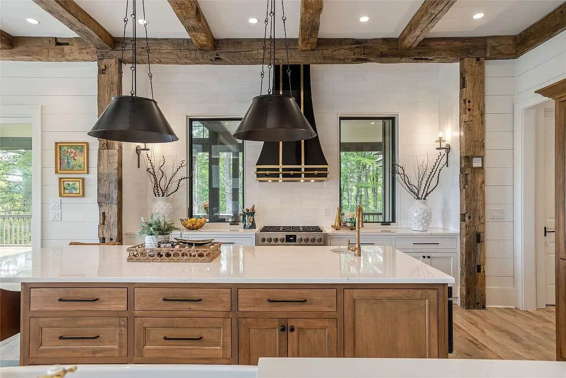 Kitchen island with warm wood cabinetry, white quartz countertop, dual black pendant lights, and black and brass range hood beyond