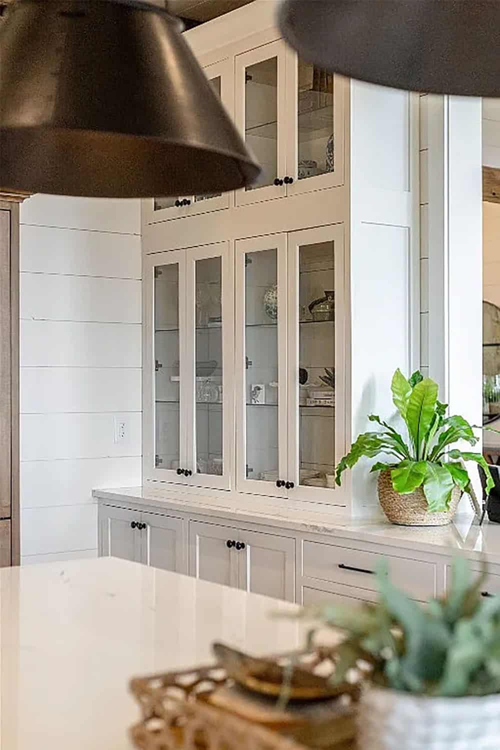 Floor-to-ceiling glass-front white kitchen cabinets with black knobs displaying china and dishware beside a quartz countertop