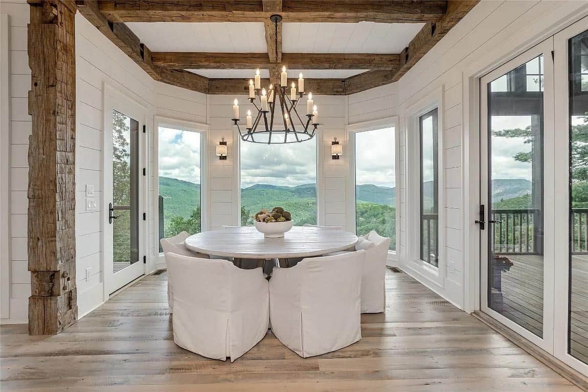 Octagonal dining nook with round table, white slipcovered chairs, iron candle chandelier, and wraparound mountain views through white shiplap walls