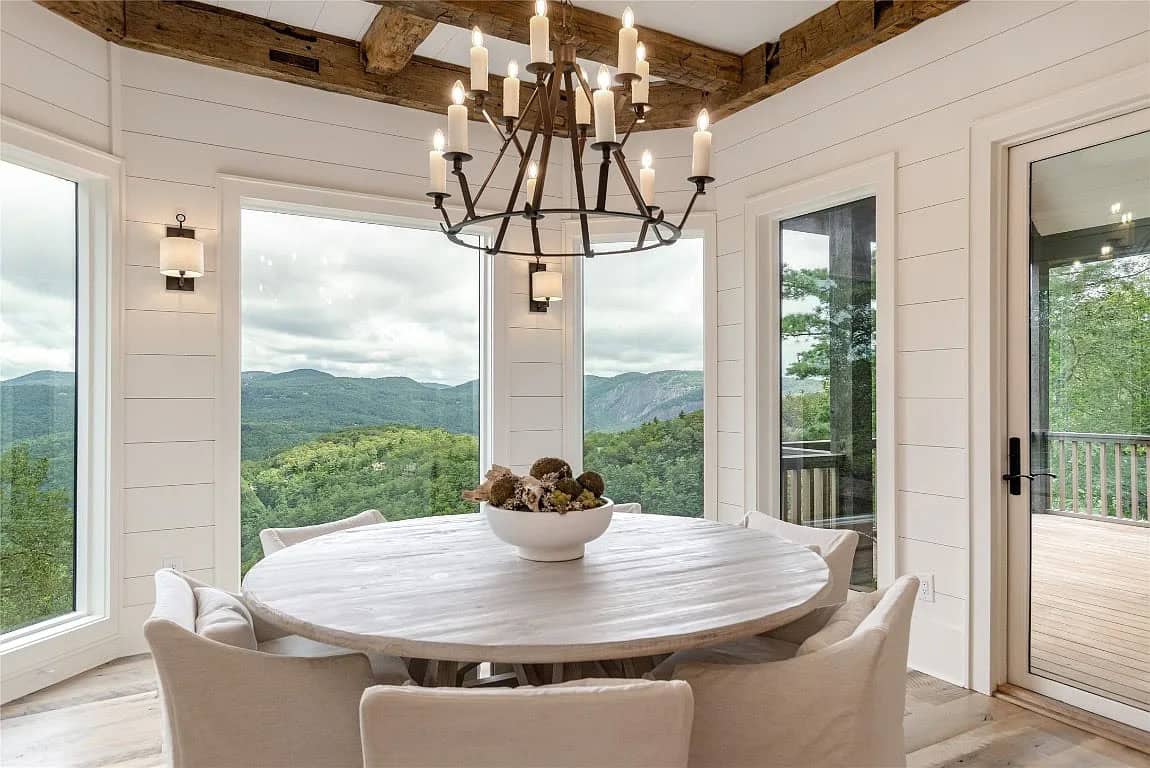 Round dining table with white linen chairs beneath iron candle chandelier framing panoramic Blue Ridge Mountain views through corner windows
