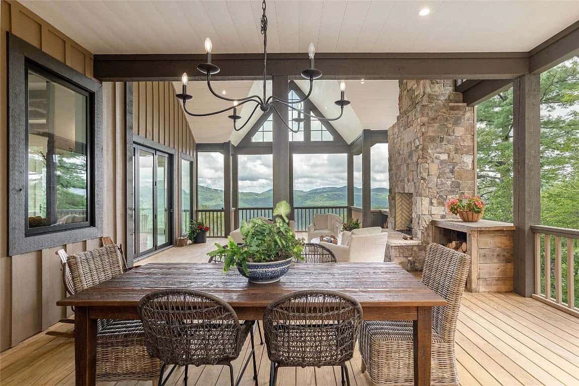 Covered porch dining area with reclaimed wood farm table, wicker chairs, iron chandelier, stone fireplace, and mountain views beyond