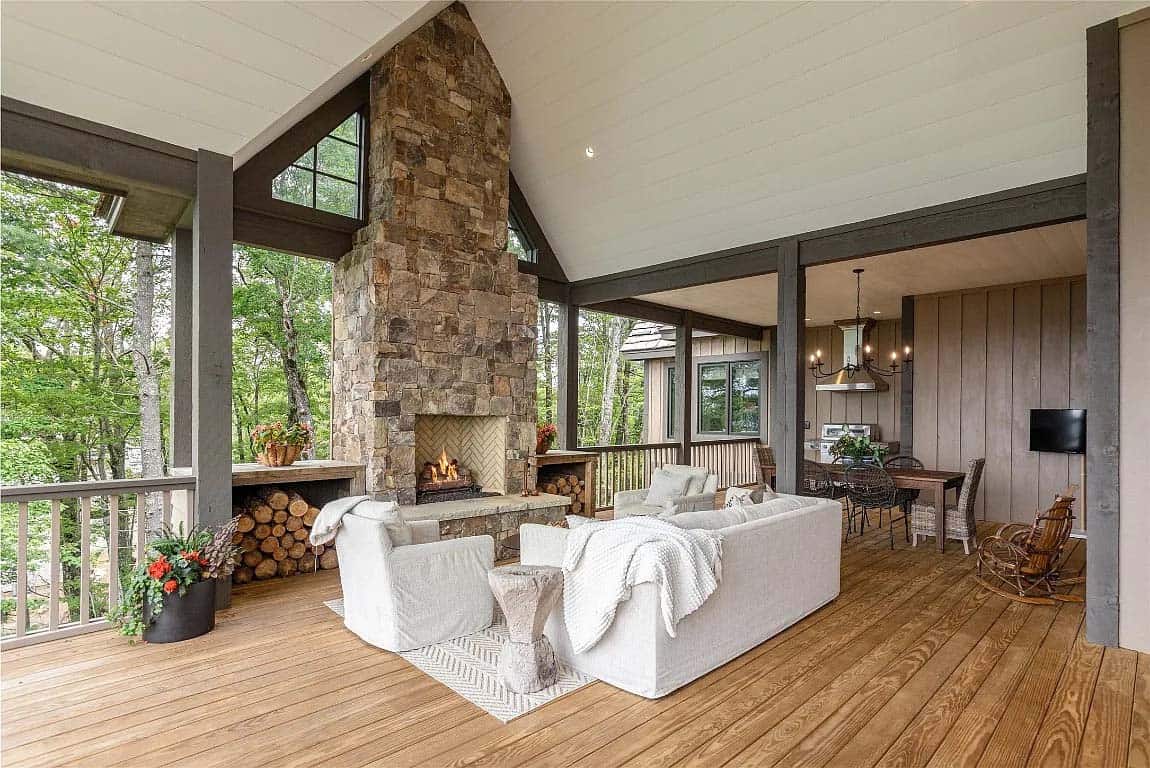 Covered porch with floor-to-ceiling stone fireplace, white slipcovered seating, stacked firewood, and wooded mountain backdrop