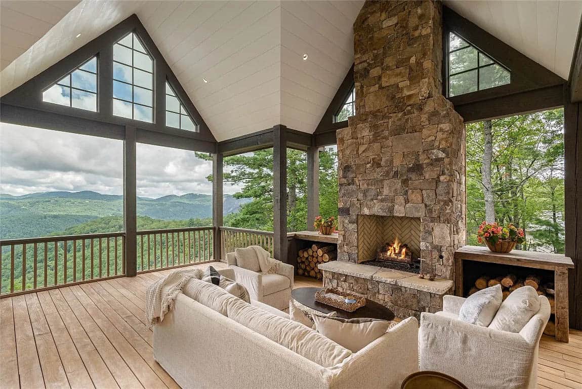 Screened porch with stone fireplace tower, cream seating, herringbone firebox with active fire, and Blue Ridge Mountain views through gable windows