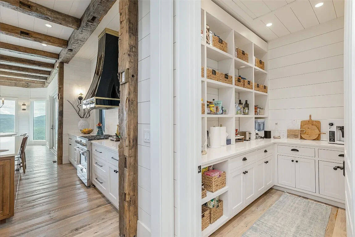 Walk-in pantry with white open shelving and wicker baskets beside view into kitchen with reclaimed beam post and black brass range hood