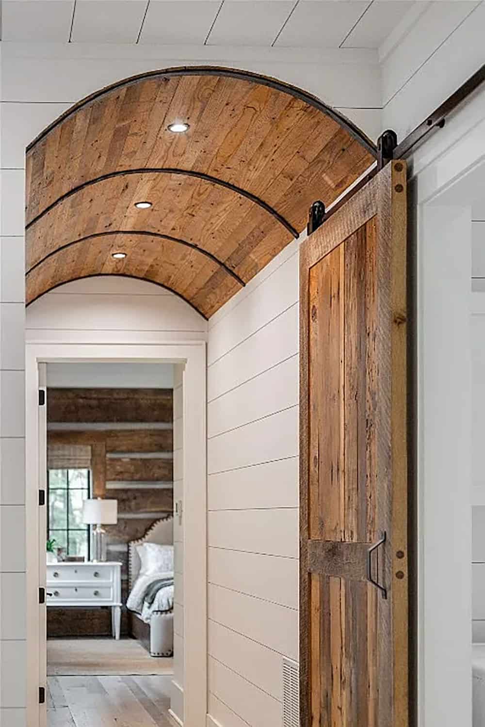Close-up of reclaimed wood sliding barn door with curved barrel-vault ceiling detail and view down hallway to bedroom with log wall