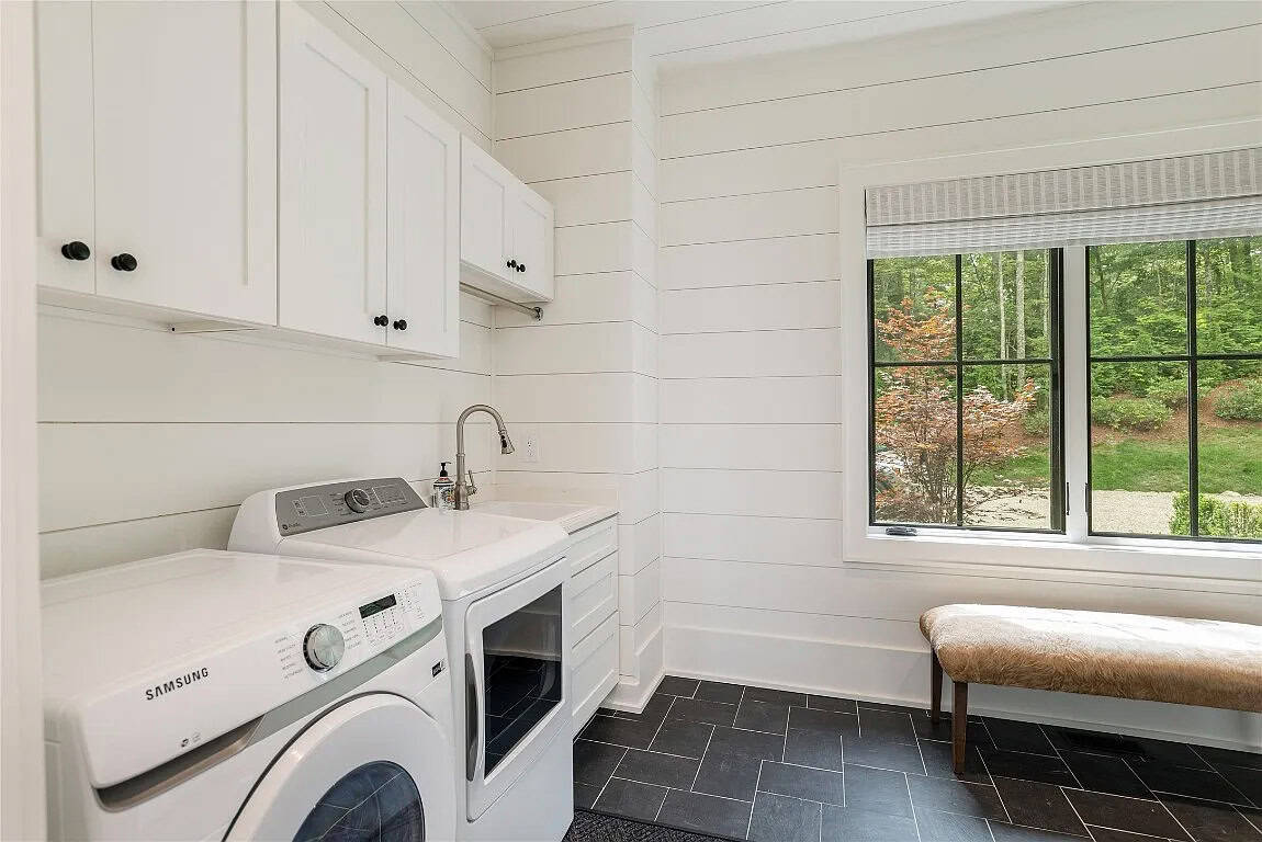 White shiplap laundry room with Samsung washer and dryer, white cabinetry, utility sink, dark tile floor, and black-framed window