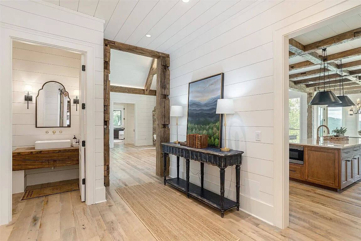Hallway junction showing powder room with vessel sink vanity, reclaimed beam doorway, console table with mountain artwork, and kitchen beyond