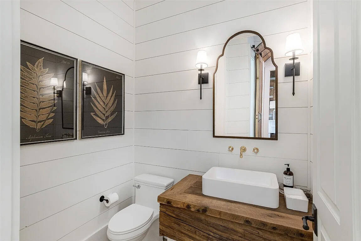Powder room with white shiplap walls, reclaimed wood floating vanity, vessel sink, brass wall faucet, and arched wood-framed mirror