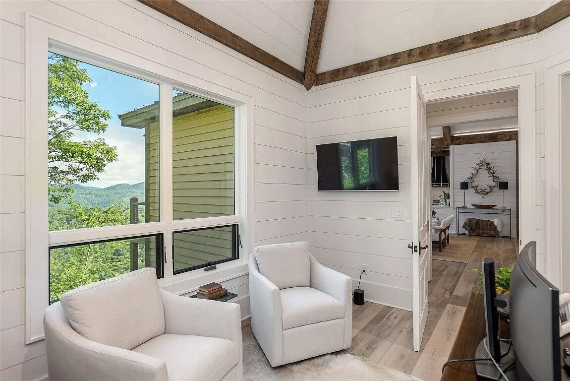 Home office sitting area with two white swivel chairs, wall-mounted TV, shiplap walls, vaulted beam ceiling, and mountain view window