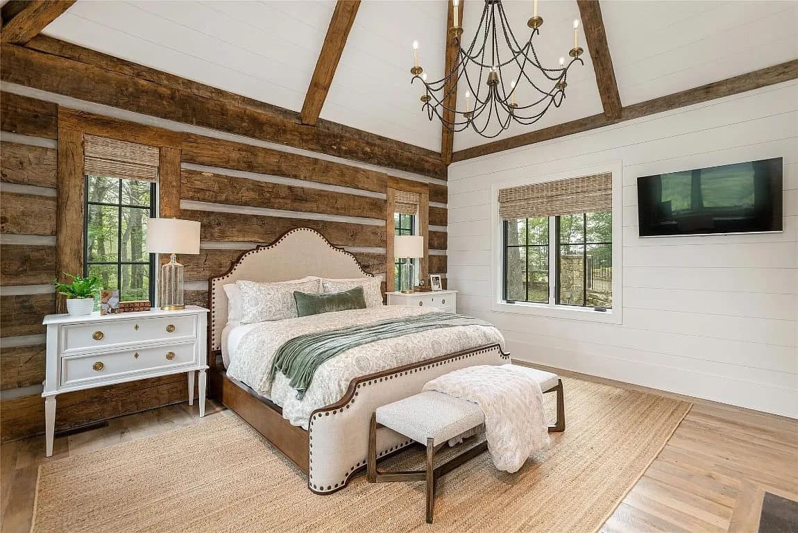 Primary bedroom with upholstered nailhead bed, log wall, vaulted shiplap ceiling, reclaimed beams, iron chandelier, and white nightstands
