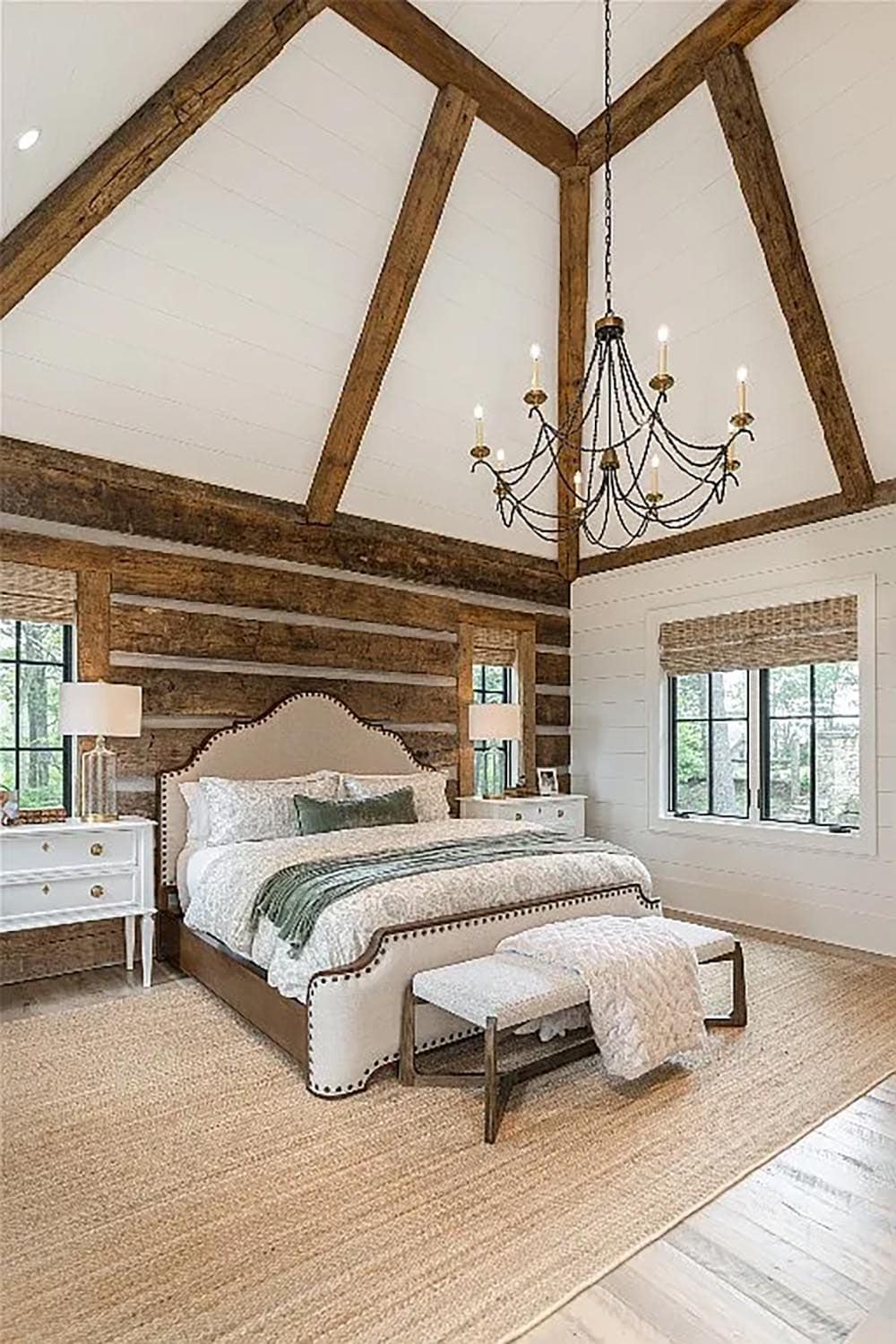 Primary bedroom showcasing soaring vaulted ceiling with reclaimed timber beams, iron swag chandelier, upholstered bed, and log accent wall