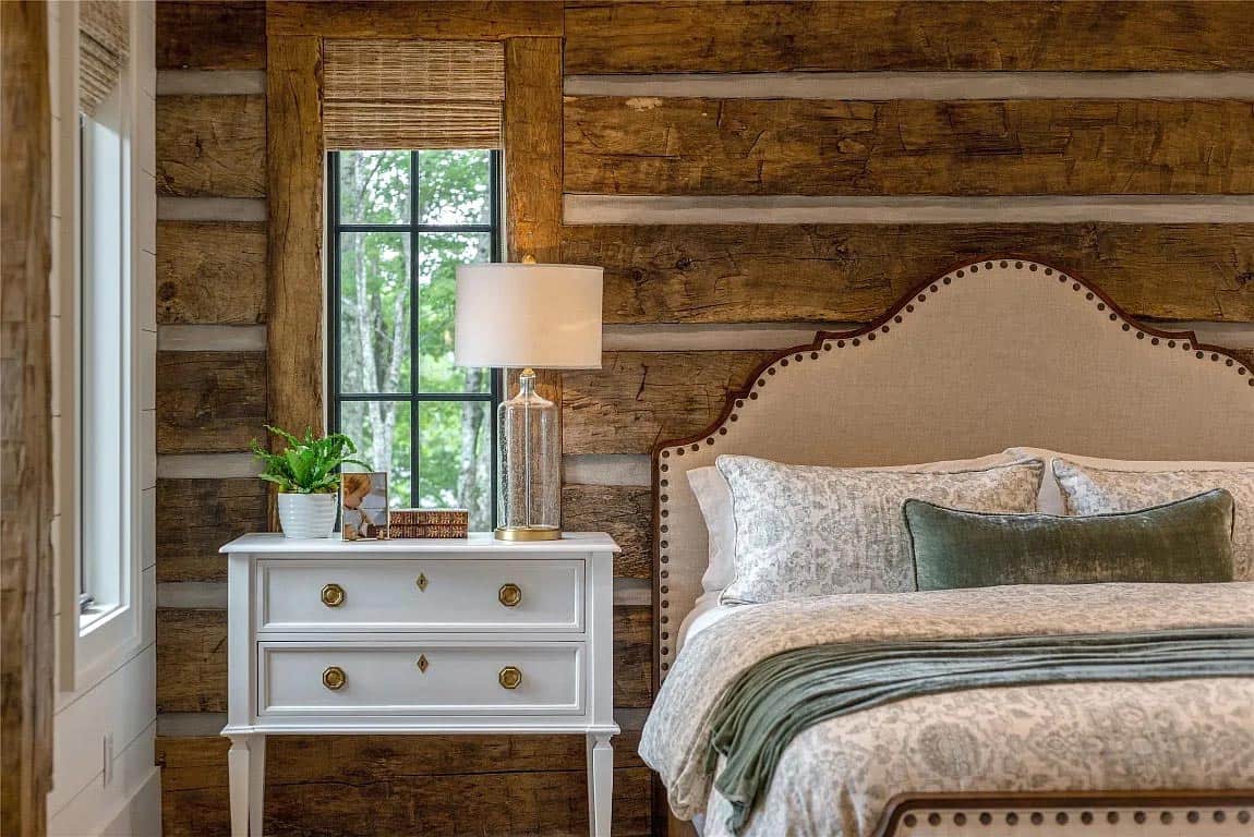 Close-up of primary bedroom nailhead upholstered headboard against rustic log wall with white nightstand and glass lamp