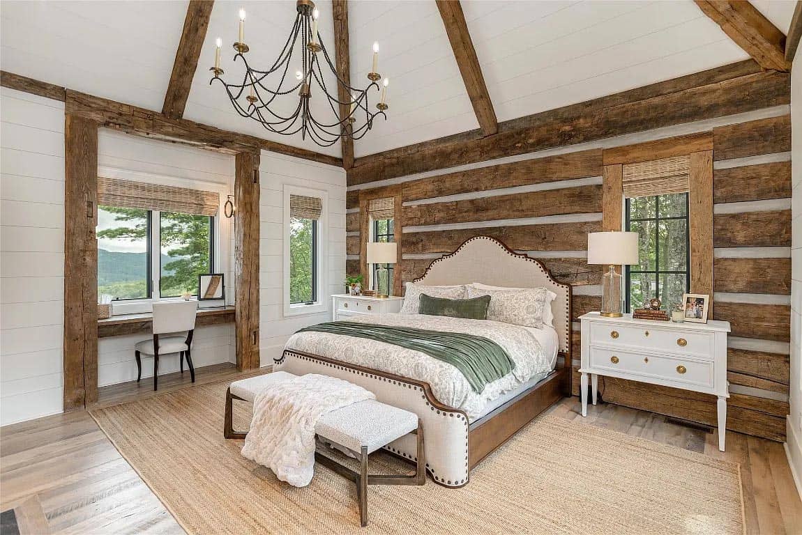 Primary bedroom with log walls, reclaimed beam vaulted ceiling, iron chandelier, upholstered bed, and built-in window desk nook beyond