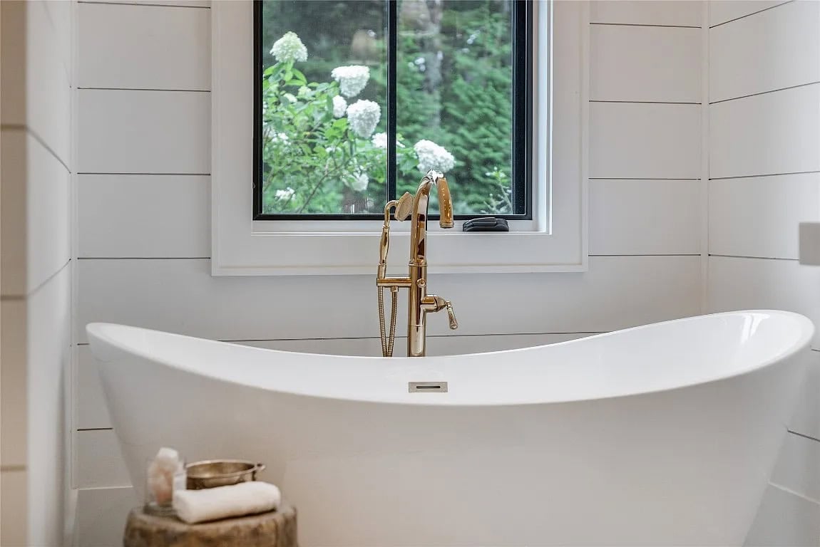 Close-up of white freestanding soaking tub with gold floor-mount faucet beside black-framed window overlooking blooming hydrangeas