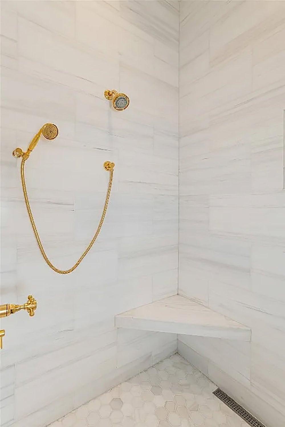 Walk-in shower interior with marble-look tile walls, corner bench, hexagon mosaic floor, and polished gold rain and hand shower fixtures