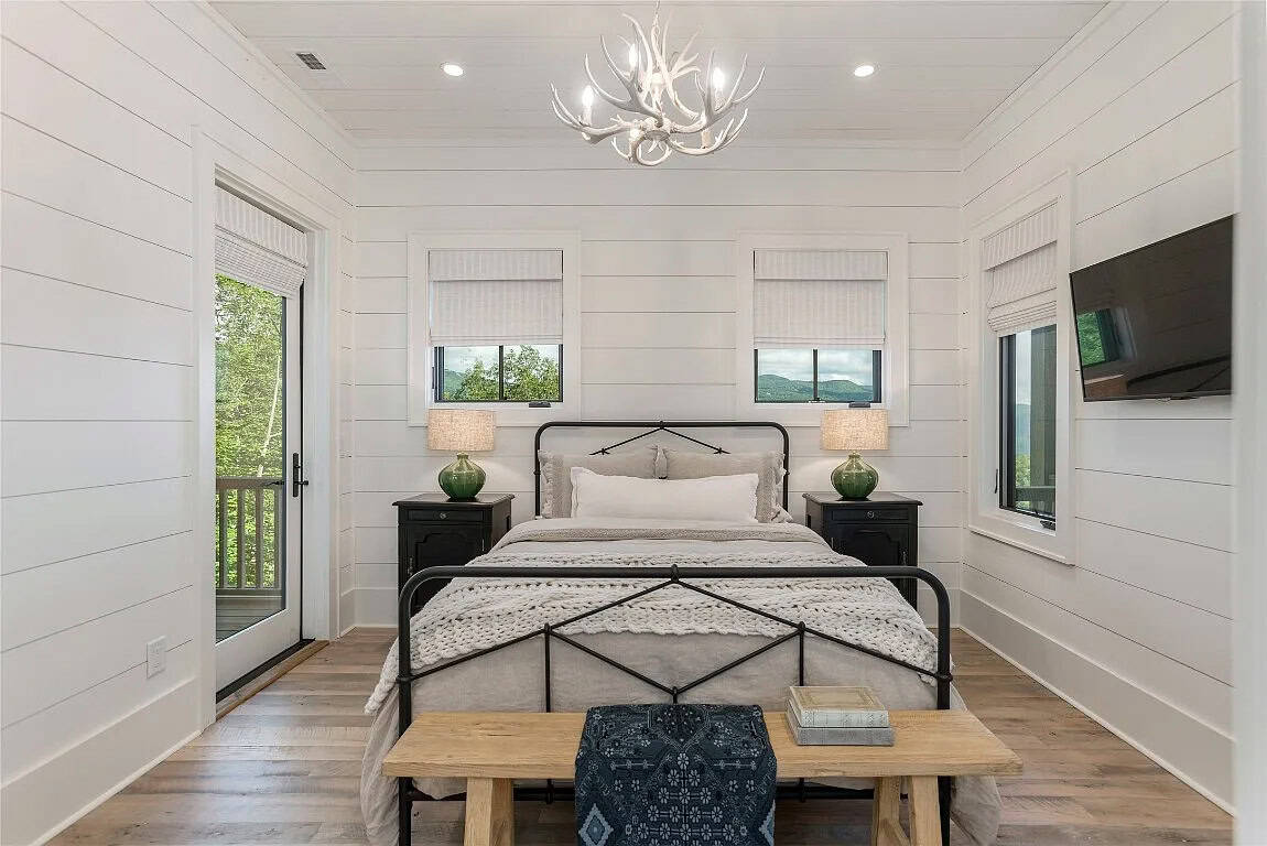 Guest bedroom viewed straight-on with black iron bed, white antler chandelier, shiplap walls, mountain-view windows, and deck door