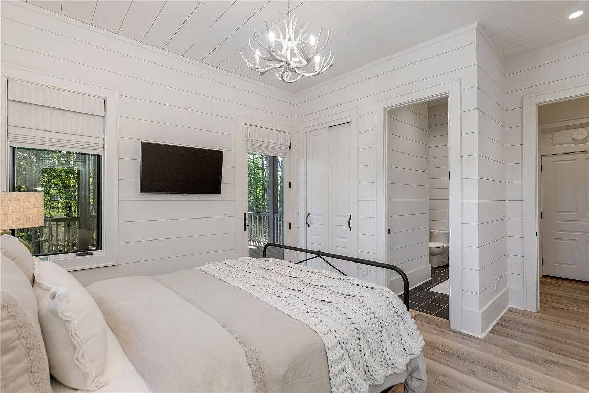 Guest bedroom with white shiplap walls, antler chandelier, black iron bed, chunky knit throw, wall-mounted TV, and deck door