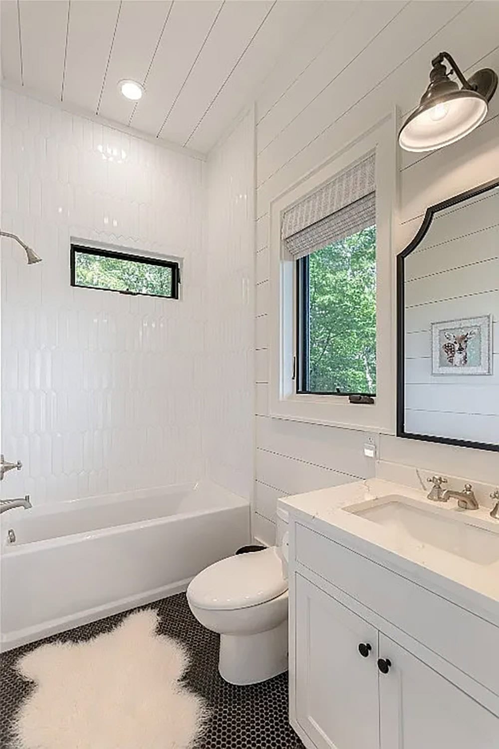 Children's bathroom with white sculpted tile tub surround, black penny tile floor, white vanity, black-framed mirror, and industrial sconce