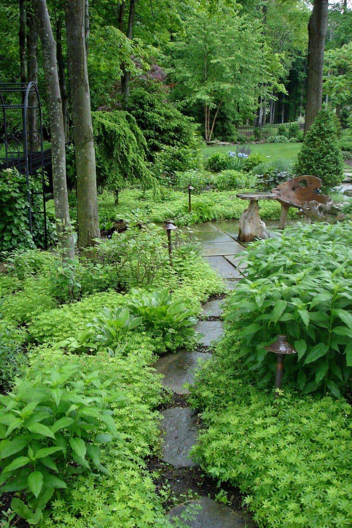 A bluestone path cuts through a lush shade garden carpeted in low-growing groundcover, with path lighting, a rustic wood stump table, and a black iron arbor among the trees