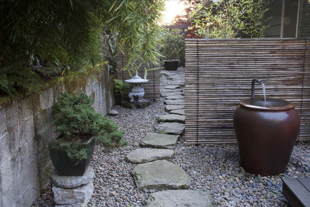 Alt tag: A Japanese-inspired shaded side yard in Eugene, Oregon, with a gravel and stepping-stone path, bamboo screens, a ceramic water feature, juniper bonsai, and a stone lantern