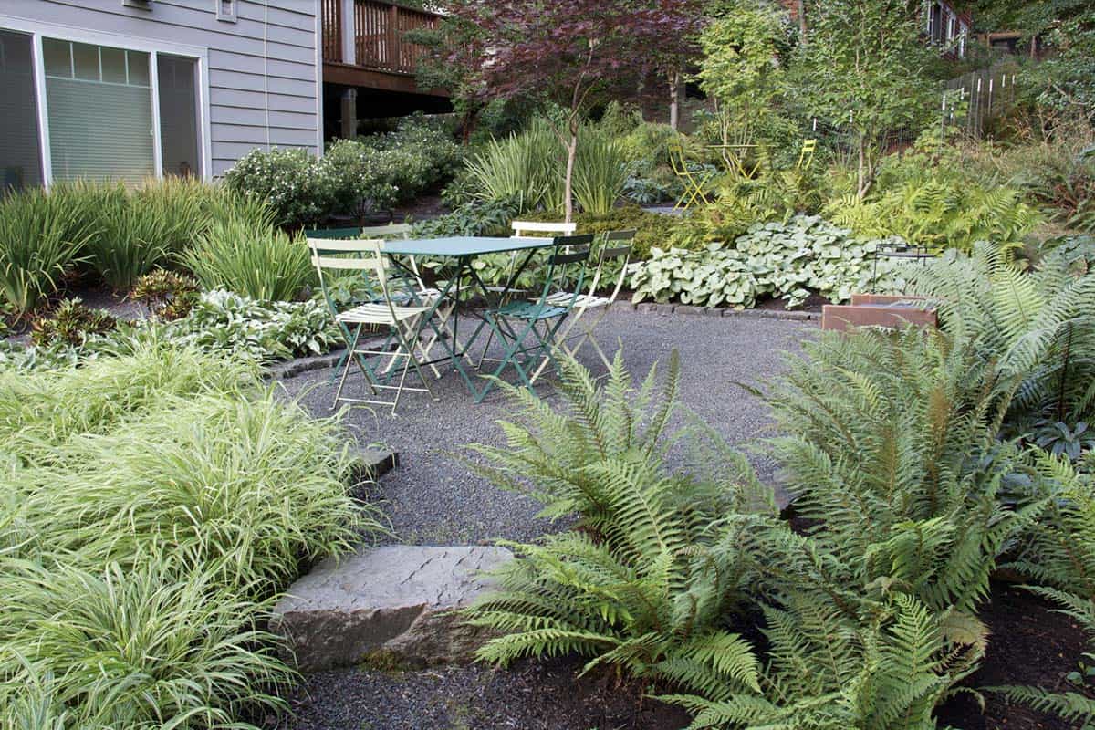 A gravel terrace with teal bistro table and chairs surrounded by ferns, hostas, and ornamental grasses in a shaded backyard garden