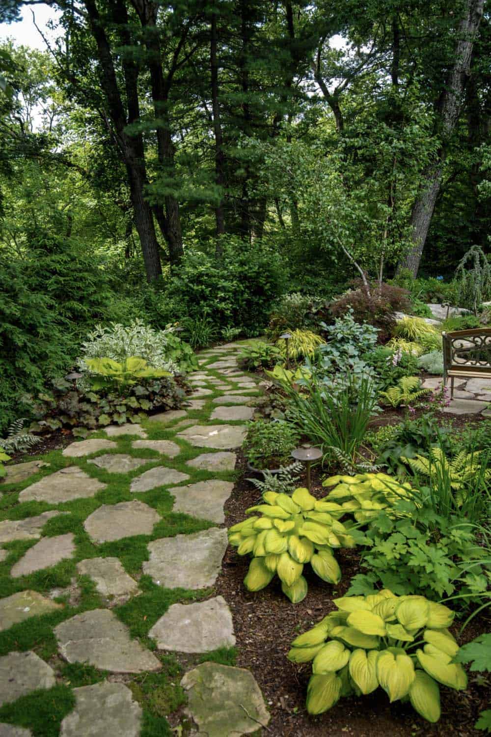 A wide irregular flagstone path set in moss winds through a lush woodland shade garden, bordered by chartreuse and variegated hostas, ferns, and a Japanese maple, with a garden bench visible at the edge