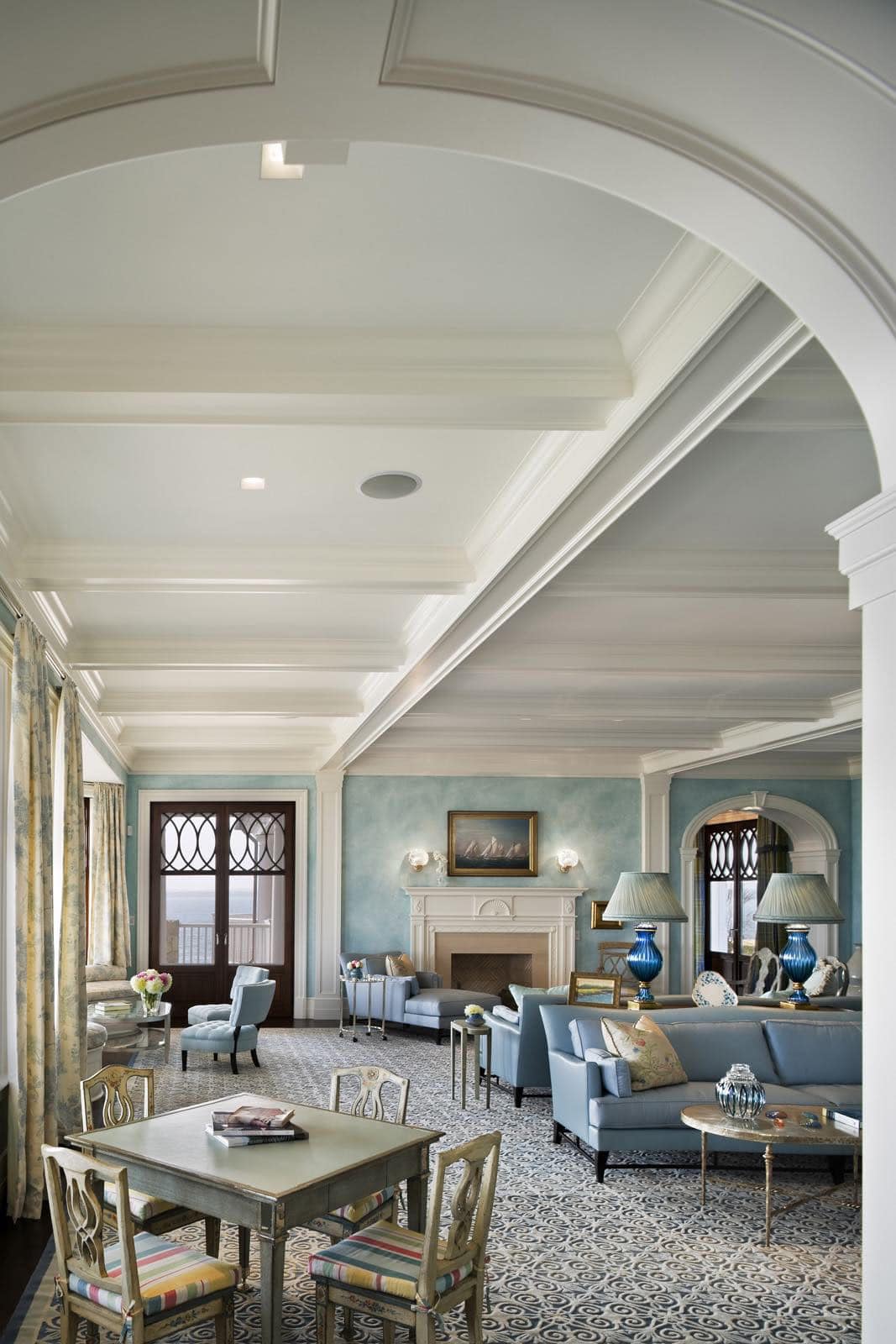 Spacious coastal living room with coffered ceiling, blue accents, fireplace, and ocean-view doors
