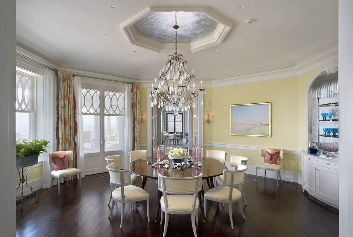 Formal dining room with octagonal tcoffered ceiling, crystal chandelier, round mahogany table, cream upholstered chairs, and yellow walls with ocean views