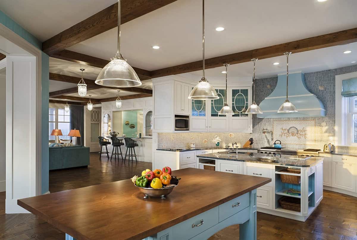 Coastal-style open kitchen with white cabinetry, blue island, exposed wood beams, glass pendant lights, and mosaic shell backsplash