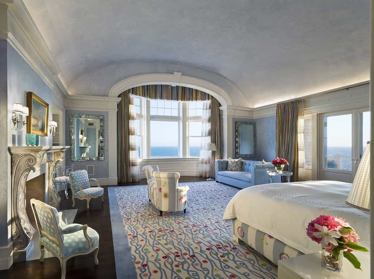 Luxurious master bedroom with barrel-vaulted ceiling, bay window with ocean view, ornate marble fireplace, and blue floral area rug