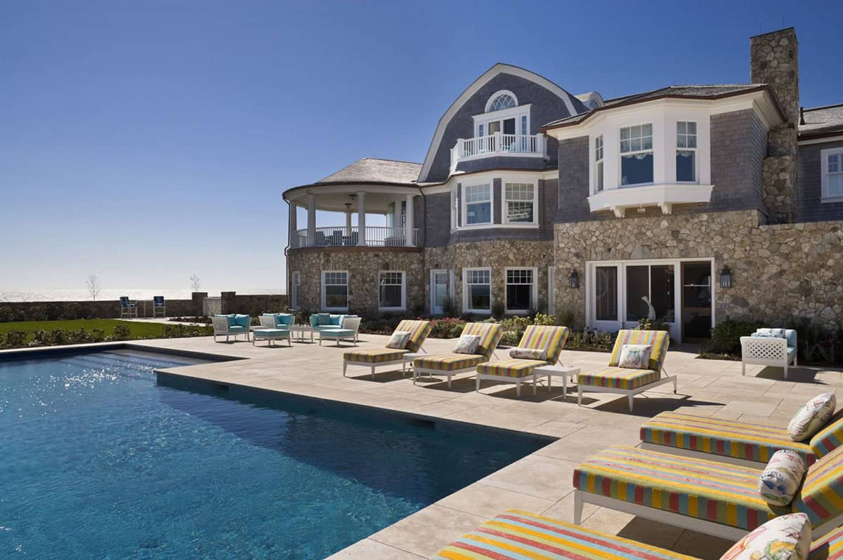Shingle-style oceanfront estate with gambrel roof, stone foundation, rectangular pool, and colorful striped lounge chairs
