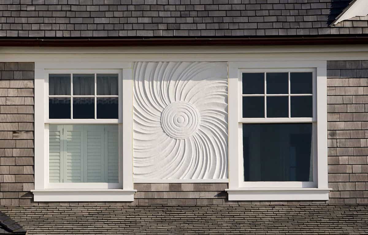 Close-up of white spiral shell relief sculpture mounted between windows on a cedar shake exterior wall