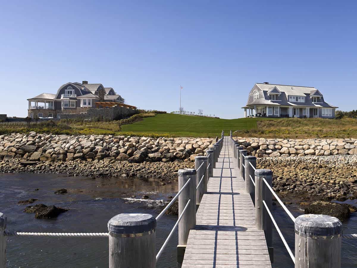 Wooden dock leading toward two shingle-style coastal homes set on a grassy oceanfront bluff