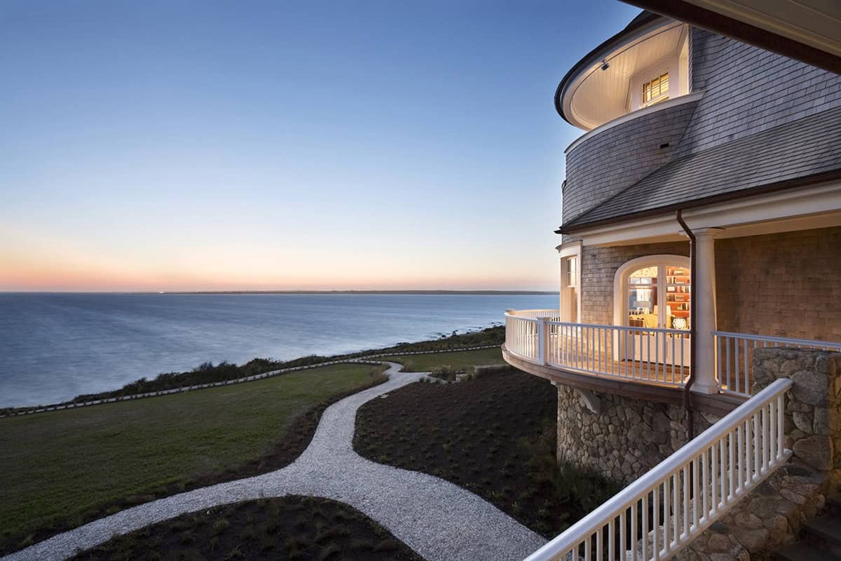 Curved shingle-style tower with illuminated wraparound porch at dusk overlooking a winding garden path and calm ocean