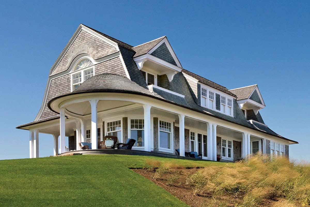 Shingle-style coastal home with sweeping gambrel roof, round covered porch with white columns, and ocean dune setting