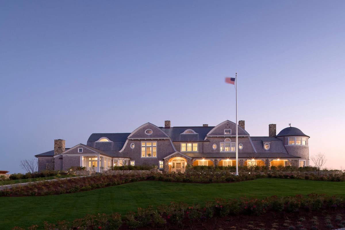 Grand shingle-style estate at twilight with glowing windows, American flag, and manicured lawn
