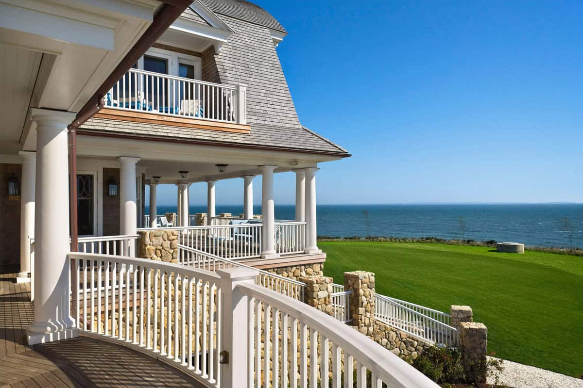  Curved oceanfront porch with white columns and railings framing a shingle-clad turret, stone walls, and sweeping ocean lawn views