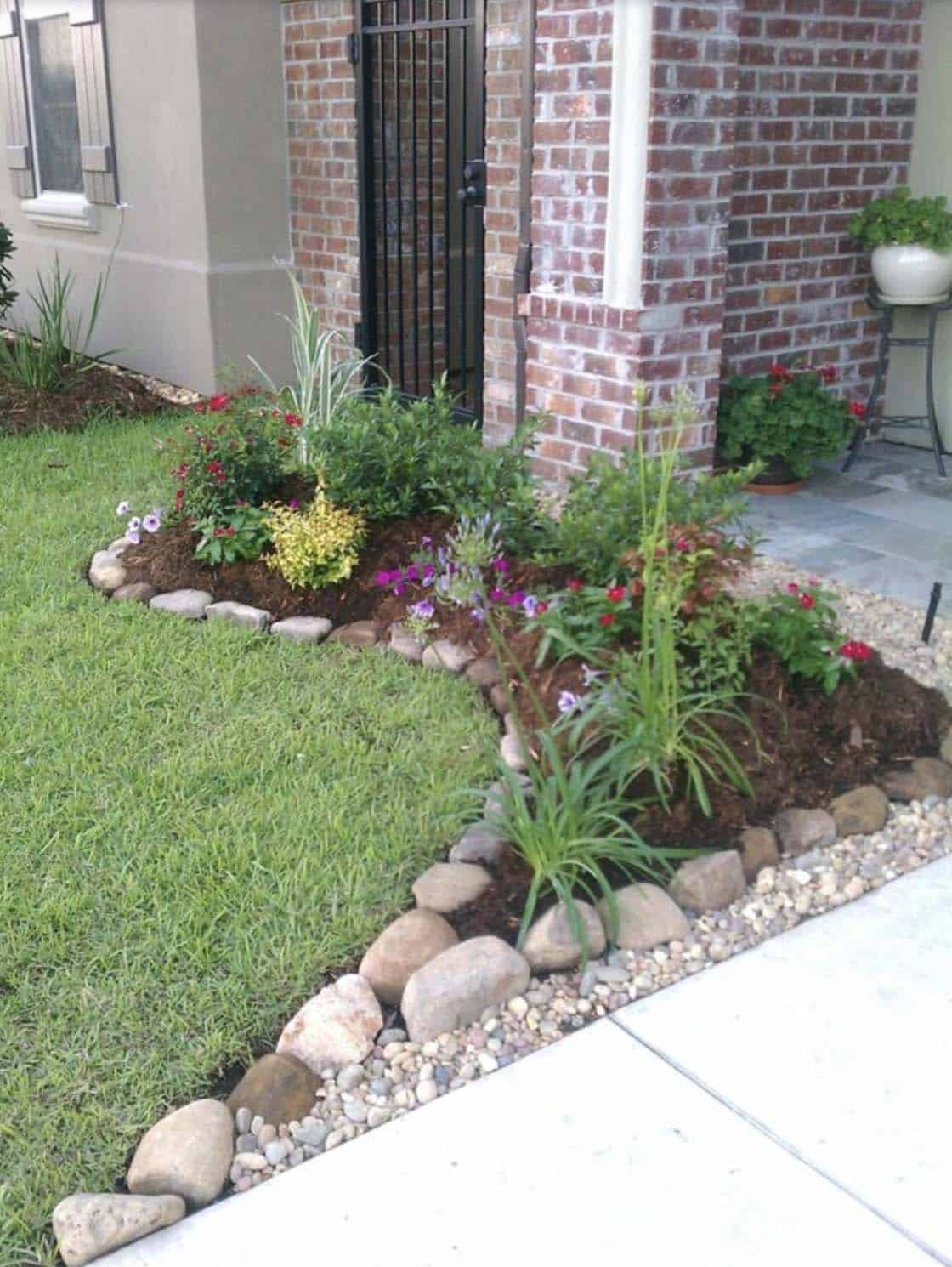 Curved river rock edging along a front garden flower bed with colorful blooms and mulch beside a brick house