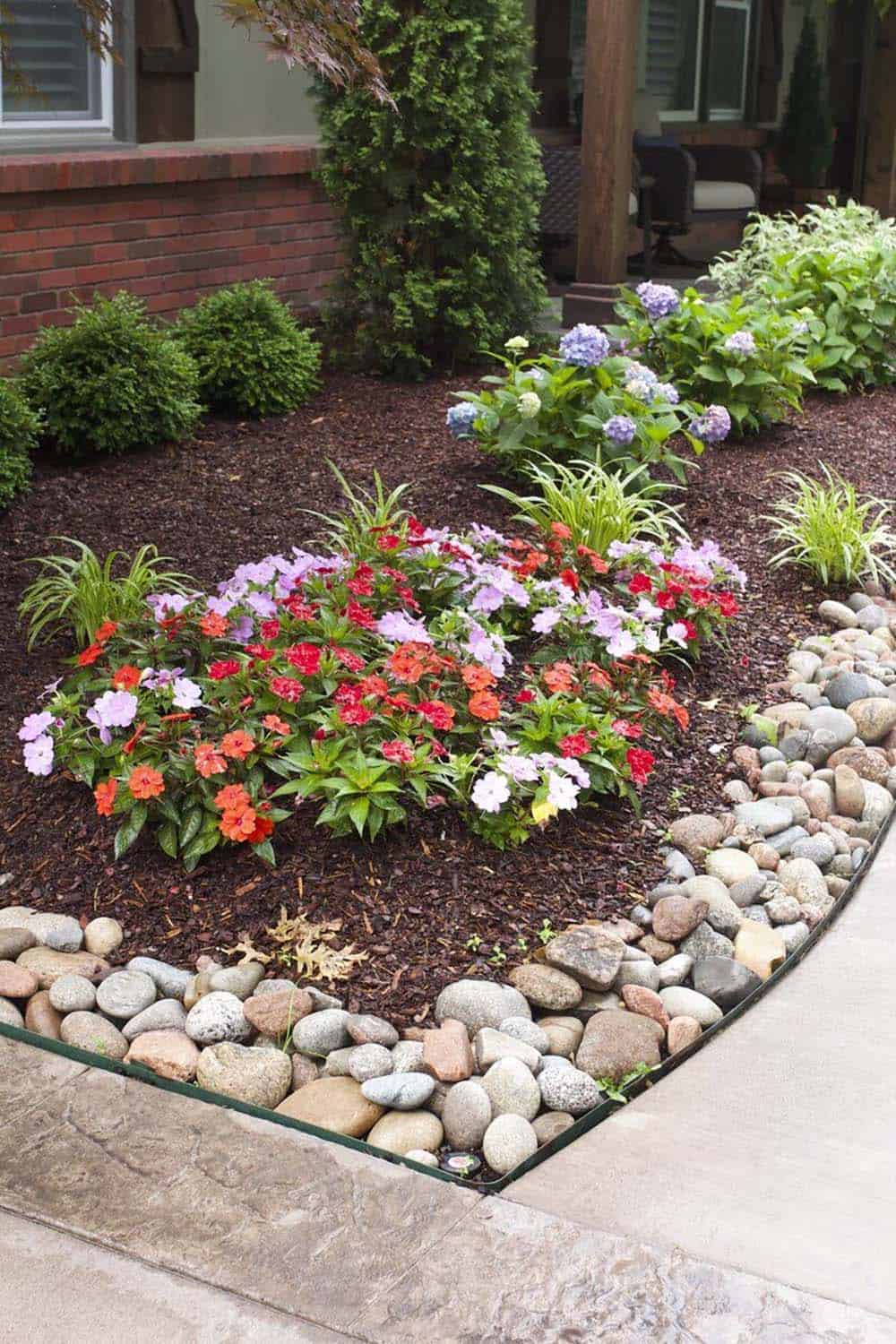River rock channel edging along a curved mulched garden bed with impatiens, hydrangeas, and boxwood beside a brick home