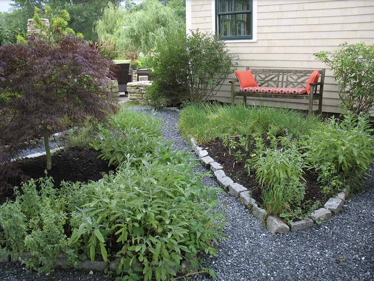 Granite cobblestone edging separating herb garden beds from a dark gravel path beside a shingle-style home