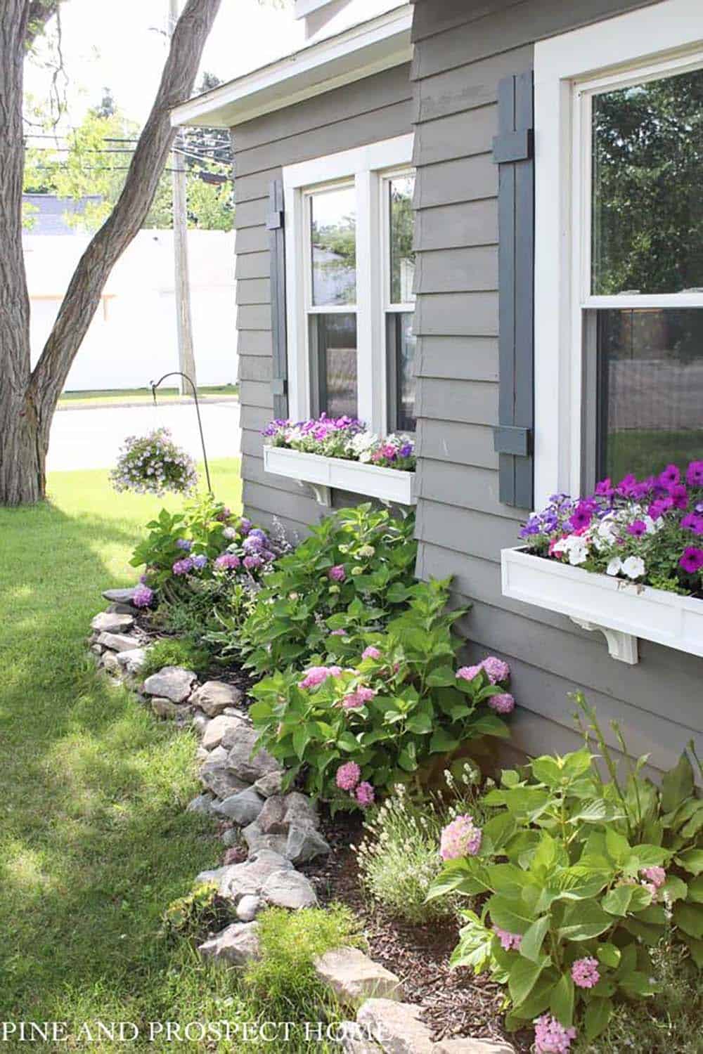 Mixed river rocks edging a cottage-style side garden bed with hydrangeas and petunias beneath window boxes on a grey home