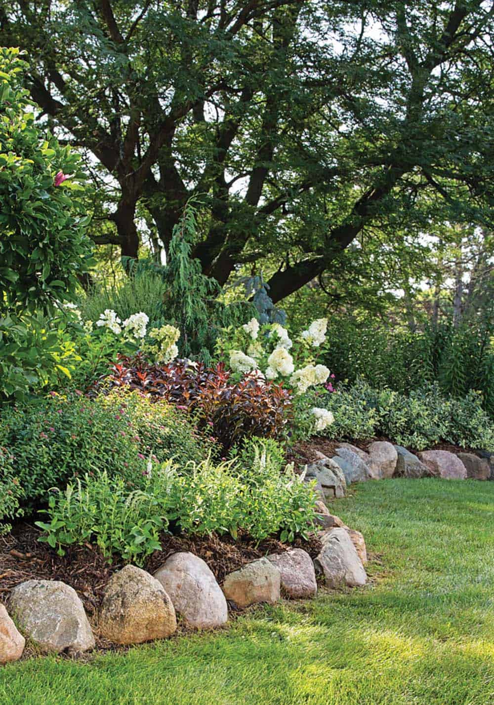 Large fieldstone boulders edging a sweeping mixed perennial border with hydrangeas and shrubs beneath mature trees