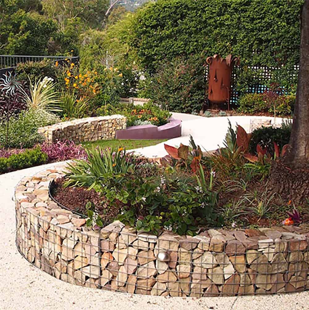 Curved gabion basket raised garden bed filled with mixed stone and tropical plantings beside a gravel path