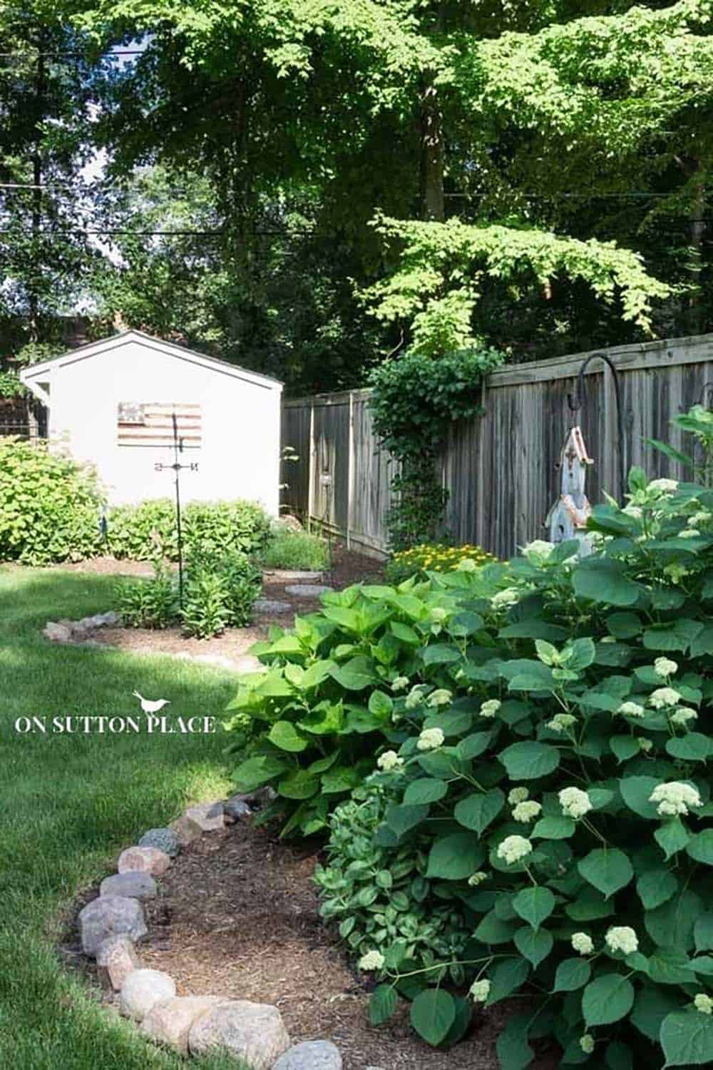 Rounded river rock edging along a curved backyard garden bed with hydrangeas and a decorative birdhouse