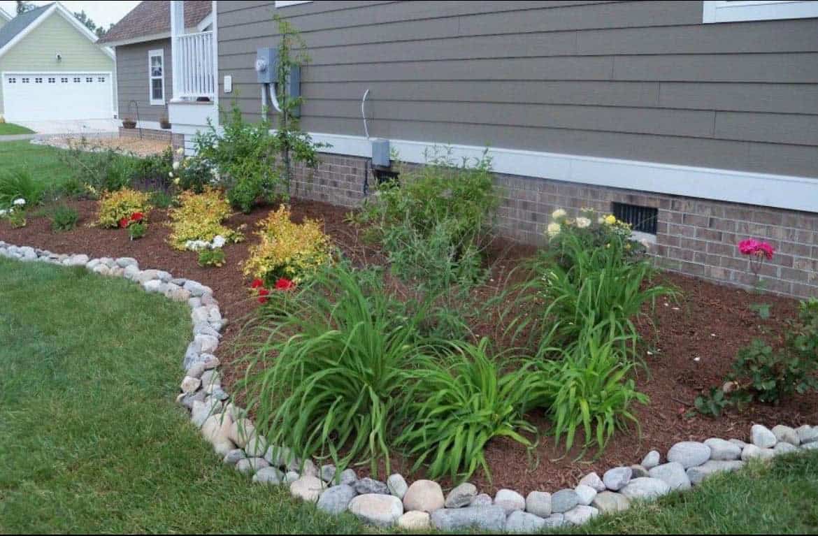 Pale river rock edging along a curved front garden bed with daylilies, peonies, and mulch beside a grey house