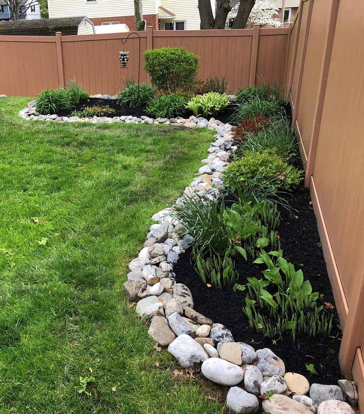 White and grey river rock edging along curved black mulch garden beds with hostas and irises in a fenced backyard