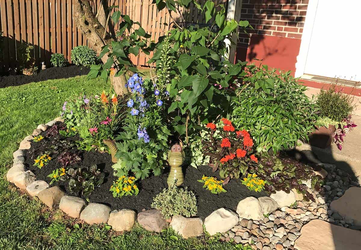 Rounded fieldstone edging around a colorful mixed annual and perennial corner garden bed beside a brick home