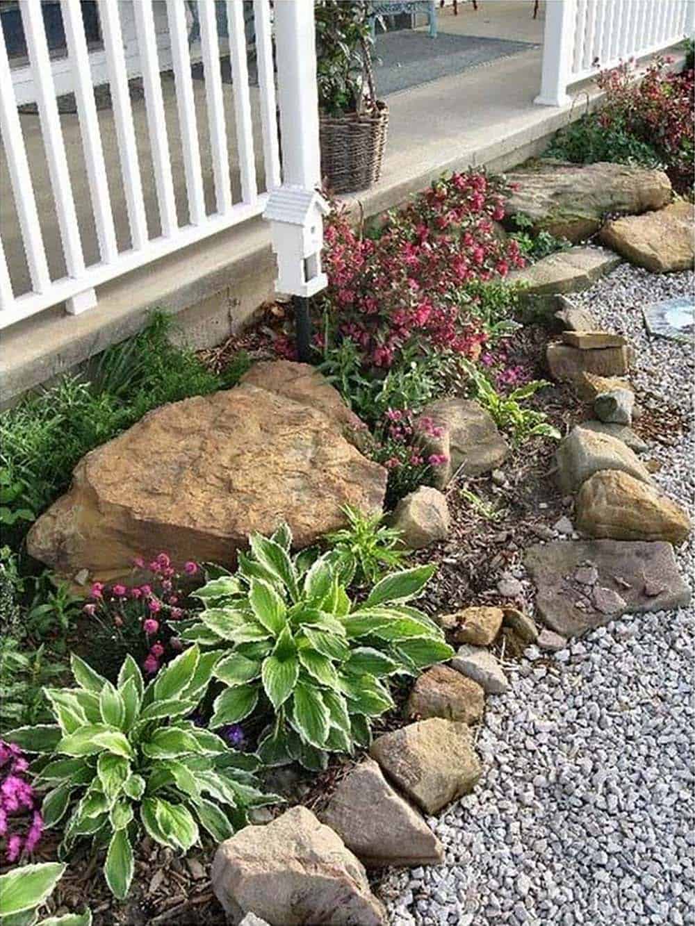 Large natural fieldstone boulders edging a cottage garden bed with hostas and pink flowers beside a white porch railing