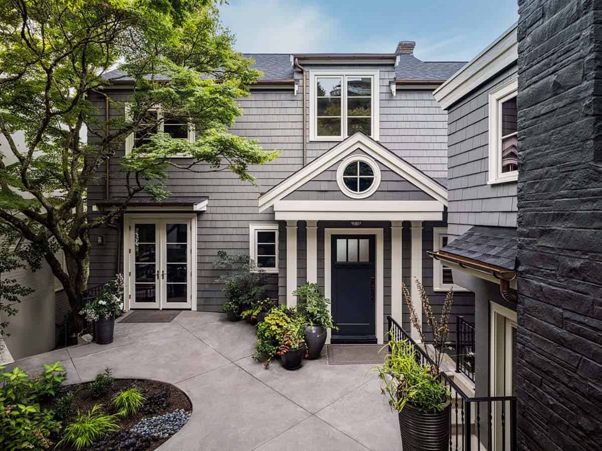 Gray shingle exterior of renovated boathouse with circular window, black front door, and lush courtyard landscaping
